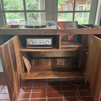 Audio Element - A wooden cabinet with two open doors reveals an audio amplifier inside, placed in a room with large windows that overlook a backyard with outdoor furniture and greenery. The cabinet is made of natural wood with visible grain patterns, and the interior contains a shelf and space for additional equipment. The room has a tiled floor and shelves with books and photographs on the right side. - Electronics Near Me - Pasadena, California