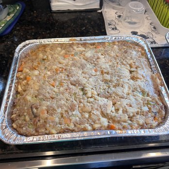 The Bagel Shoppe - Super moist and flavorful stuffing - A large aluminum tray filled with cooked stuffing containing visible pieces of carrots, celery, and bread crumbs is positioned on a dark granite kitchen counter. The background includes various kitchen items such as a dish rack with napkins, plastic containers, and utensils, indicating a home kitchen setting. - Caterers Near Me - Fishkill, New York