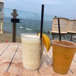 two drinks on a table overlooking the ocean - Two drinks are positioned on a wooden table outdoors, with a view of the ocean and sandy beach in the background. One drink appears to be a creamy or frozen beverage with a lemon wedge garnish, while the other is a darker, possibly iced tea or cocktail. The setting includes outdoor chairs stacked against a wall and a glass railing separating the seating area from the beach. The sky is partly cloudy.