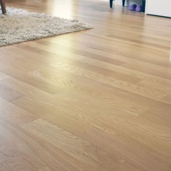 Summit Hardwood Floors - The image depicts a section of a residential interior with a hardwood floor featuring a natural wood finish. The floorboards are arranged in a parallel pattern, showing wood grain and texture. A portion of a plush, light-colored rug and furniture legs are visible in the background, along with a doorway and some household items. The lighting suggests natural daylight illuminating the space. - Flooring Near Me - Hanson, Massachusetts