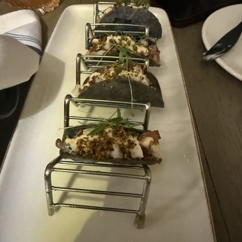 Ingrano - Octopus tacos. Yum! - A white rectangular plate holds three octopus tacos, each topped with herbs and garnishes. The tacos are presented with metal taco holders, and the plate is placed on a dark wooden table. Surrounding items include a napkin, a glass of beverage, and a smartphone, indicating a dining setting. - Bakeries Near Me - Annapolis, Maryland