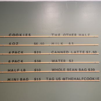 The Half Cookie - Prices! - The image shows a menu board on a plain wall, listing prices for various cookie options, beverages, and bags at The Half Cookie in Boston. The menu includes items such as 6 cookies for $6.50, a 4-pack for $25, and a mini bag for $15, with additional details about milk, canned latte, water, and bags. The board is organized with horizontal wooden slats separating the menu items, and the text is in black capital letters. - Desserts Near Me - Boston, Massachusetts