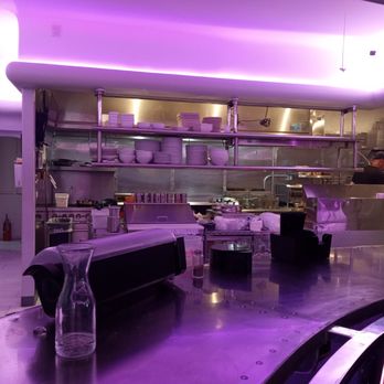 Bisbee Royale - Open kitchen - The image depicts an open kitchen in a restaurant with purple ambient lighting. Shelves above the counter hold stacks of plates and bowls. A chef is visible working in the background, and various kitchen equipment and utensils are present. The foreground features a curved table with a glass pitcher and other items. The overall setting suggests a modern dining environment with a focus on visual aesthetics. - Music Venues Near Me - Bisbee, Arizona