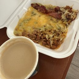 Cafe con Leche - A foam cup of coffee and a white takeout container with baked beans, cheese, shredded cabbage, and bacon on a wooden table.
