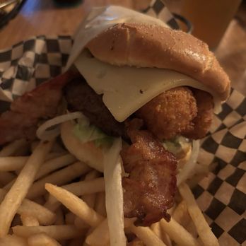 Wildfangs Grill House - Mozz Burger - The image shows a burger with fried chicken, a slice of cheese, lettuce, and bacon, topped with a bun. The burger is placed on checkered paper, accompanied by a side of French fries. In the background, there are drinks and a wooden table surface, indicating a casual dining setting. - Barbeque Near Me - Springfield, Oregon