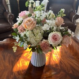 White Campanula, Peach Carnations, Seeded Eucalyptus, White Hydrangea, Orange Spray Rose, Shimmer Rose, Ruscus - A bouquet of roses, hydrangeas, lilies, and other flowers in a white vase is placed on a polished wooden table, with leather chairs and a fireplace in the background.