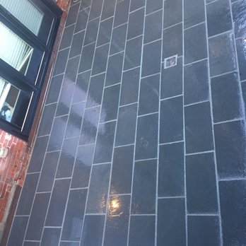 Back Yard Living - New patio pavers - The image shows a section of a patio with dark gray rectangular pavers arranged in a staggered pattern. A metal drain is embedded in the paving stones. To the left, there is a brick wall with a window that has blinds. The surface of the pavers appears wet, reflecting light, indicating recent cleaning or rain. An electrical conduit and a small pipe are visible near the brick wall, suggesting outdoor utility connections. - General Contractors Near Me - Abington, Massachusetts