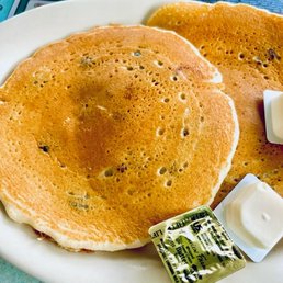 a plate of pancakes - Two golden-brown pancakes with small dark spots are presented on a white plate, accompanied by butter and syrup packets.