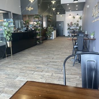 Sucre Table - Inside - The image shows the interior of a coffee shop with a modern minimalist design. There are several small tables with black chairs along the right wall, decorated with potted plants. The counter area on the left has a black finish, with shelves above holding more plants and coffee equipment. The ceiling is black with exposed lighting fixtures, and the floor is wood-look tile. A sign reading 'Coffee' is visible on the wall near the counter. - Bakeries Near Me - Tampa, Florida