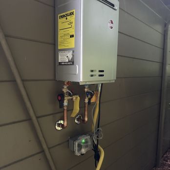 Big Chunks Plumbing - Tankless water heater - A tankless water heater is installed on the exterior of a building, mounted on a wall with visible copper plumbing connections, shut-off valves, and electrical wiring. The unit has an energy guide label indicating energy efficiency and cost. A small electrical box with a green indicator light is also attached below the heater. The wall is made of horizontal siding, and the ground below shows some debris. - Plumbing Near Me - Eureka, California
