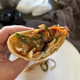 K-breakfast Burrito - A close-up of a partially eaten Korean breakfast burrito filled with shredded vegetables, scrambled eggs, and kimchi, held in a person's hand. The burrito shows a mix of colorful ingredients including green, orange, and purple vegetables. The background includes a white plate with some remaining filling, a brown cloth, and various objects on a table, indicating a casual dining setting.