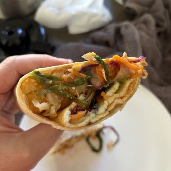 Sonbul - Korean breakfast burrito sans meat. - A close-up of a partially eaten Korean breakfast burrito filled with shredded vegetables, scrambled eggs, and kimchi, held in a person's hand. The burrito shows a mix of colorful ingredients including green, orange, and purple vegetables. The background includes a white plate with some remaining filling, a brown cloth, and various objects on a table, indicating a casual dining setting. - Comfort Food Near Me - Brooklyn, New York