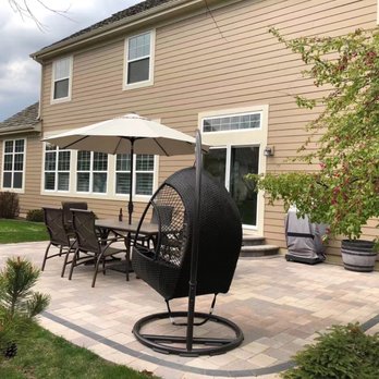 SAR Landscaping Contractors - a patio with patio furniture - The image shows a backyard patio area with a paved surface, including a dining table with six chairs and a large umbrella for shade. A black hanging chair is positioned nearby, and a potted tree with pink flowers is on the right side. The beige house in the background has multiple windows, a sliding glass door, and a small set of steps leading to the door. The scene is outdoors with neighboring houses visible in the background. - Gardeners Near Me - Elgin, Illinois