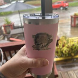 Got myself a new coffee cup while I was there. - A pink insulated tumbler with a straw, featuring the Mars Brew House logo, held by a person inside a cafe on a rainy day.