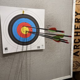 Super Fun and professionally taught! - Three arrows are embedded in a paper archery target, with two in the red zone and one near the center, mounted on a gray wall. The target has concentric circles in white, black, blue, red, and yellow. The arrows have green and red fletching, and the target is part of an indoor archery range setting.
