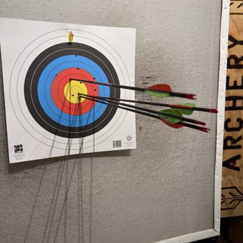 Gotham Archery - Super Fun and professionally taught! - Three arrows are embedded in a paper archery target, with two in the red zone and one near the center, mounted on a gray wall. The target has concentric circles in white, black, blue, red, and yellow. The arrows have green and red fletching, and the target is part of an indoor archery range setting. - Archery Near Me - Brooklyn, New York