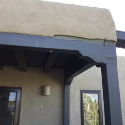 a close up of the roof of a house - The photo depicts a building corner featuring a concrete ledge with visible damage and cracks, supported by painted wooden beams. The building's exterior wall is textured, and there are windows and a light fixture visible. The scene suggests structural wear or damage to the concrete ledge and support structure.
