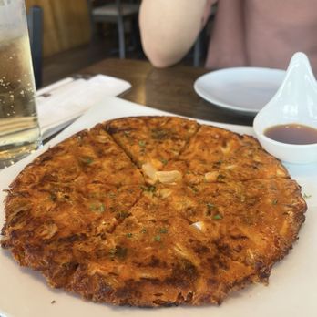 Woori - Seafood pancake - A seafood pancake, golden-brown and crispy, is placed on a white plate. It appears to contain seafood and vegetables, garnished with herbs. A small dish of dipping sauce is positioned next to the pancake. The setting includes a glass of beverage, possibly soda or sparkling water, and a person in a pink shirt is partially visible in the background, seated at a wooden table in a casual dining environment. - Korean Near Me - Arlington, Massachusetts