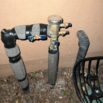 Union Plumbing - An outdoor plumbing pipe system with insulated pipes, valves, and a pressure gauge, situated near a wall and on gravel ground. - Plumbing Near Me