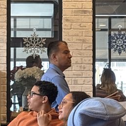 Manager that thinks he is better than anyone else in the place. - The image shows an indoor restaurant scene with a man in a blue shirt standing near a brick wall with two windows decorated with snowflake ornaments. In the foreground, two women are seated at a table; one woman wears glasses and an orange hoodie, and the other has glasses and a gray beanie. The background includes reflections of other patrons and interior decor, indicating a casual dining environment.