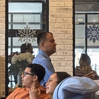 Maxfield's Pancake House - Manager that thinks he is better than anyone else in the place. - The image shows an indoor restaurant scene with a man in a blue shirt standing near a brick wall with two windows decorated with snowflake ornaments. In the foreground, two women are seated at a table; one woman wears glasses and an orange hoodie, and the other has glasses and a gray beanie. The background includes reflections of other patrons and interior decor, indicating a casual dining environment. - Breakfast & Brunch Near Me - Milwaukee, Wisconsin