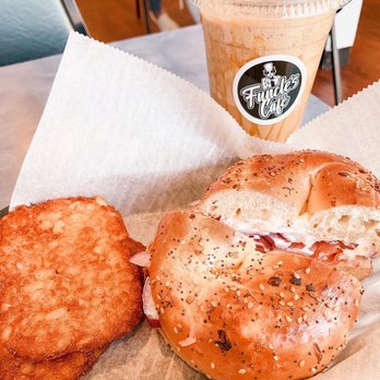 Funcle's Café - Bagel - The image shows a breakfast meal on a tray with a bagel sandwich filled with cream cheese and meat, a crispy hash brown, and an iced coffee in a clear plastic cup with a straw, featuring the Fundle's Cafe logo. The tray is lined with parchment paper, and the background suggests a casual dining setting. - Coffee & Tea Near Me - Hamden, Connecticut