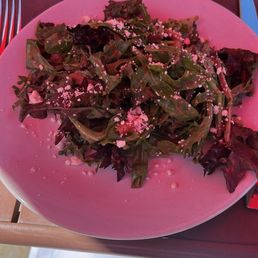 Salad Pasta & Salad or Soup - A plate of mixed leafy greens salad featuring different types of lettuce and greens, topped with pink crumbles, possibly cheese or seasoning. The salad is served on a pink plate, with a glass of beverage and utensils visible on a pink outdoor table. The setting appears to be an outdoor dining area with natural lighting.