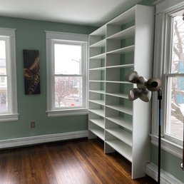 Bedroom - A white, tall bookshelf with multiple shelves is placed against a green wall in a room with hardwood flooring and two windows.