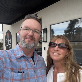 El Verde Inn - Just arrived after a long leg of our road trip. - A man and woman are taking a selfie in front of a building with a sign that reads "El Verde Inn." The man has short hair, glasses, and a beard, and is wearing a plaid shirt. The woman has long hair, sunglasses, and is wearing a white top. The background shows windows, a wall-mounted light, and trees reflected in the windows. The setting appears to be outdoors during daytime. - Hotels Near Me - Albuquerque, New Mexico