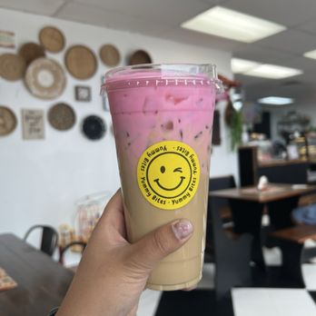 Yummy Bites - A person holds a large plastic cup filled with a layered iced drink, with pink on top and beige below, inside a cafe. The cup has a yellow smiley face sticker with a wink and the text 'Yummy Bites.' The background shows a cafe interior with wall decor, tables, chairs, and ceiling lights, indicating a casual dining environment. - Desserts Near Me - Amarillo, Texas