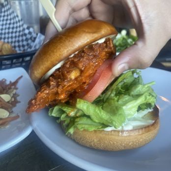 The Kosmos - Fried Chicken Sandwich - A fried chicken sandwich with lettuce, tomato, and sauce is held by a hand over a white plate with additional food in the background. - Bars Near Me - Albuquerque, New Mexico
