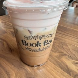 Mocha with raspberry cold foam - A large iced coffee with a layer of raspberry cold foam on top is served in a clear plastic cup labeled 'Book Bar.' The drink contains ice cubes and appears to be a coffee beverage with milk or cream. The cup is on a wooden table, with a cafe environment visible in the background, including people seated and engaged in activities.