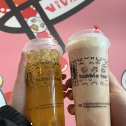 Vivi Signature Fruit Tea, Hot Taro Creme Brulee Milk Tea - Two bubble tea drinks are shown being held up in front of a pink wall decorated with bubble tea branding and graphics. The left drink is a yellowish fruit tea with visible fruit pieces, while the right drink appears to be a creamy milk tea. Both cups have clear lids and are branded with the bubble tea company's logo and contact information.