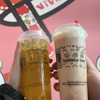 Vivi Bubble Tea - Vivi Signature Fruit Tea, Hot Taro Creme Brulee Milk Tea - Two bubble tea drinks are shown being held up in front of a pink wall decorated with bubble tea branding and graphics. The left drink is a yellowish fruit tea with visible fruit pieces, while the right drink appears to be a creamy milk tea. Both cups have clear lids and are branded with the bubble tea company's logo and contact information. - Bubble Tea Near Me - Columbus, Ohio