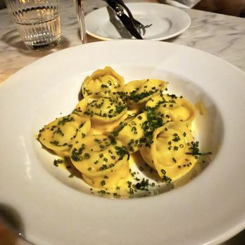 The Snail - Crab pasta - A white, round plate contains several pieces of ravioli pasta, garnished with chopped green herbs. The dish is placed on a marble table, with a glass of white wine, a glass of water, and a plate with cutlery visible in the background. The setting appears to be a restaurant, with a person partially visible in the background wearing a light-colored shirt. - American Near Me - Brooklyn, New York