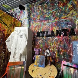 The image shows an art studio with walls covered in multicolored splatter paint and abstract patterns. A white protective suit hangs on a folding screen, and a small round table holds two stuffed animal sculptures and a small canvas painting. Chairs with paint-streaked surfaces are positioned around the table. The studio features a sign spelling 'ATLANTA' on the wall, indicating the location. The space is filled with creative, expressive artwork and supplies.