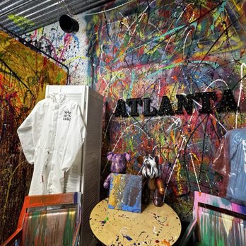 The Splatter Studio - The image shows an art studio with walls covered in multicolored splatter paint and abstract patterns. A white protective suit hangs on a folding screen, and a small round table holds two stuffed animal sculptures and a small canvas painting. Chairs with paint-streaked surfaces are positioned around the table. The studio features a sign spelling 'ATLANTA' on the wall, indicating the location. The space is filled with creative, expressive artwork and supplies. - Team Building Activities Near Me - Atlanta, Georgia