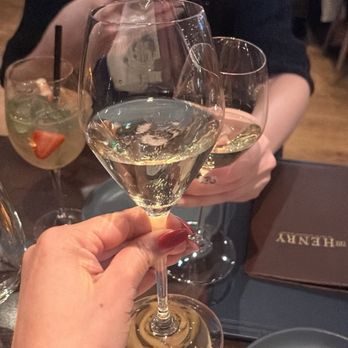 The Henry - Champagne - A person holds a glass of champagne in a restaurant setting, with other drinks, food, and a woman using a phone visible in the background. - New American Near Me - Phoenix, Arizona