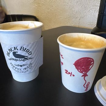 Blackbird Coffee House - Two coffee cups with hot beverages are placed on a black table at Black Bird Coffee House in Albuquerque, NM, with one cup featuring a logo and the other a red balloon drawing. - Coffee & Tea Near Me - Albuquerque, New Mexico