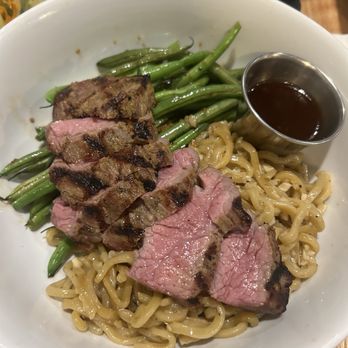 Rawbar - Garlic steak bowl with noodles - A bowl containing sliced grilled steak, green beans, and ramen noodles, with a small cup of dark sauce on the side. - Sushi Bars Near Me - Chico, California