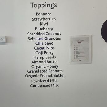Deep Purpl - Toppings - A list of toppings such as bananas, strawberries, kiwi, blueberry, shredded coconut, selected granolas, chia seed, cacau nibs, goji berry, hemp seeds, almond butter, organic honey, granulated peanuts, organic peanut butter, powdered milk, and condensed milk is displayed on a white wall. The list is printed in black text and appears to be part of a menu or product options in a retail or food service setting. - Juice Bars & Smoothies Near Me - Chicago, Illinois