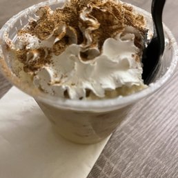 Tres Leches - A cold coffee drink in a clear plastic cup, topped with whipped cream and sprinkled with cocoa powder. A black straw is inserted into the drink, and the cup is placed on a white napkin on a wooden table surface. The background is blurred, focusing on the beverage.