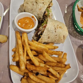 El Huarache - Shrimp Burrito - A rectangular white plate holds two burritos, French fries, and a small cup of orange sauce, with a restaurant setting. - Mexican Near Me - Queens, New York