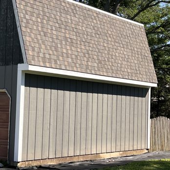 Wolf Development - The image shows a building with a steeply sloped roof covered in brown asphalt shingles. The exterior walls are clad in vertical gray siding, and there is a white trim along the edge of the roof. A wooden fence is visible on the right side, and the building is set against a background of trees with green foliage. The ground in front of the building is covered with black landscaping fabric and grass. - Roofing Near Me - Hoffman Estates, Illinois