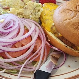 Le Fritter - A ceramic plate holds a cheeseburger with a toasted bun, melted cheese, and a beef patty. Accompanying the burger are sliced red onions, tomato slices, and a serving of potato salad with visible potato chunks and mayonnaise. The plate is placed on a wooden surface, with a knife partially under the burger.