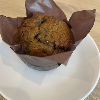 Sproutz - Brickell - Muffin - Blueberry - A blueberry muffin with visible blueberries is presented in a brown paper wrapper, sitting on a plain white plate. The muffin appears freshly baked with a slightly cracked top, and the setting includes a wooden surface background. - Juice Bars & Smoothies Near Me - Miami, Florida