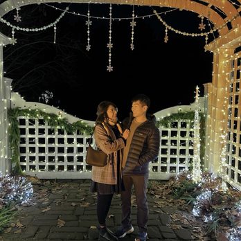 Newfields - A woman and a man stand together under festive holiday lights and decorations at night, with a white lattice fence and greenery in the background. - Parks Near Me - Indianapolis, Indiana