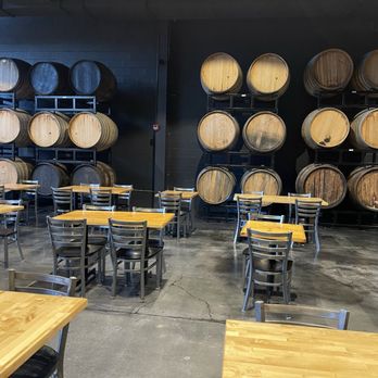 Trillium - Canton - Multiple wooden barrels are stacked on black metal racks against a black wall in an industrial-style space. The barrels vary in size and color, with some appearing darker or more weathered. In the foreground, there are several wooden tables with metal chairs arranged around them, suggesting a tasting room or brewery seating area. The floor is concrete with visible cracks, and the overall environment has a modern, industrial aesthetic. - Breweries Near Me - Canton, Massachusetts
