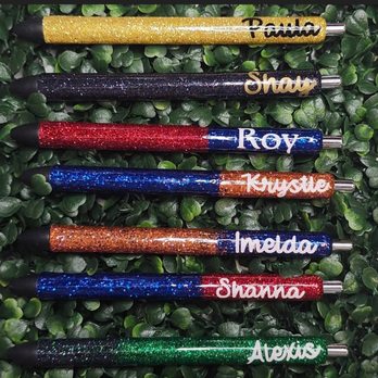 Pretty Pens - Six glitter pens with various colors, including gold, black, red, blue, orange, and green, are laid out on a green leafy background. Each pen features a metallic tip and a name written in a contrasting color: Paula, Shay, Roy, Krystie, Imelda, and Shanna. The pens are decorated with glitter, making them visually striking and suitable for personalized stationery or gifts. - Customized Merchandise Near Me - Austin, Texas