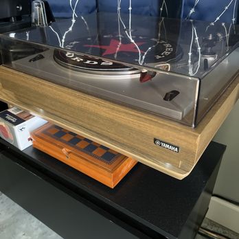 The Sound Guy - Yamaha YP 701 - A Yamaha YP-701 vintage turntable with a clear dust cover is positioned on a black table. The turntable features a wooden base and a black platter with a red and black label. Underneath the table, there is an orange chessboard and a small box. The background includes a blue curtain with white star-like patterns. - Electronics Repair Near Me - Phoenix, Arizona